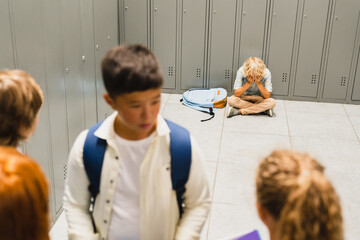Close up focused lonely sad schoolboy crying at the school hall floor while his classmates ignoring...