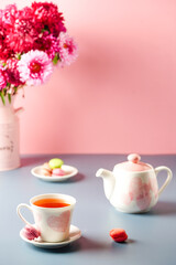 white cup with pink hearts a teapot, sweets and a bouquet of flowers on a pink-gray background. Breakfast, morning tea.