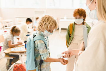 Teacher measuring temperature her students classmates schoolchildren before lesson wearing protective medicine face masks against coronavirus Covid19 due to lockdown restrictions.