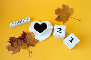 Calendar for September 21 : the name of the month in English, cubes with the number 21,a white heart-shaped cup on a saucer with tea, maple leaves, yellow background, top view