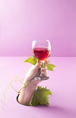 A hand holding a glass of red wine with a vine leaf on a purple background. Autumn minimal concept.