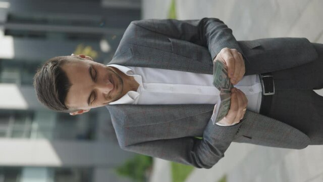 Happy good looking man in business style holding dollars and calculating. Young Handsome Rich Man Wearing Stylish Suit Counting Money Standing in the street near Office Building Vertical Video