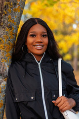 Portrait of a beautiful african american young woman with a smile in a fashionable black casual jacket near a tree in a park with bright golden autumn foliage