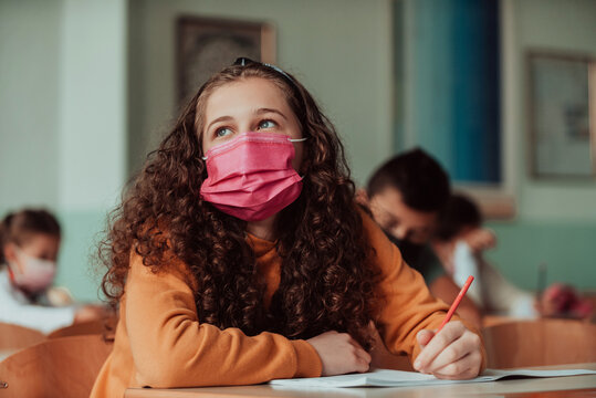 Caucasian Kid School Girl Wearing Face Mask Studying In Classroom Or At Home Sitting At Desk. Mixed Race Child Doing Homework Learning Online. Social Distance Remote Education Concept