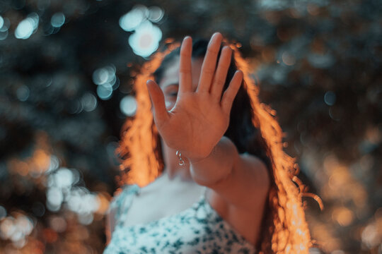Mujer joven de pelo rizado evitando una fotografia haciendo la se&ntilde;al de parar o stop junto a la naturaleza de los arboles y la puesta de sol por la ma&ntilde;ana