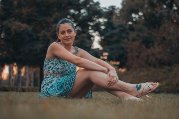 Chica joven de vestido azul relajándose sentada en el césped de un parque junto a los arboles y...