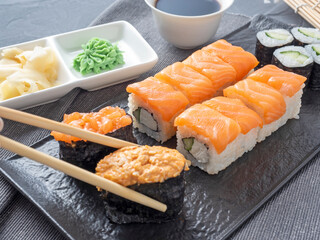 a variety of Japanese rolls and sushi on a textured plate. Side view. bamboo sticks of ginger and sauce nearby.