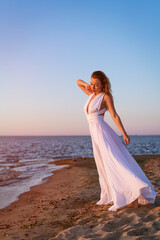 Fototapeta na wymiar a young, beautiful, adult brunette woman in a white fluttering dress stands on the sandy shore of the bay. Take a full-length portrait at sunset
