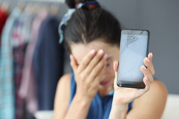 Sad woman covering her eyes with hand and holding broken mobile phone closeup