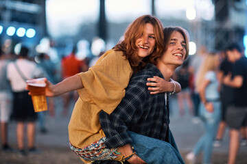A happy smiling hippie couple having a piggyback ride at a rock concert. The girl is holding a plastic glass of fresh beer while the man is holding her.