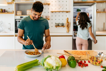 A young, happy couple prepares dinner together at the weekend. Man is stirring vegetables in the...