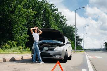 A young girl stands near a broken-down car in the middle of the highway and looks under the hood....