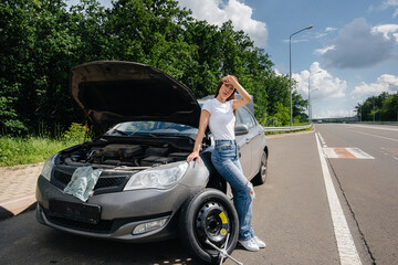 A young girl stands near a broken car with a broken wheel in the middle of the highway and is frustrated waiting for help on a hot day. Breakdown and breakdown of the car. Waiting for help.