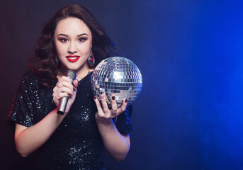 Naklejka premium lifestyle and people concept: woman wearing black dress, holding disco ball and singing into microphone
