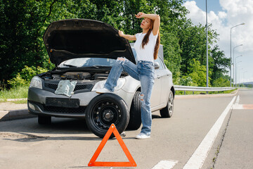 A young girl stands near a broken car in the middle of the highway and tries to change a broken wheel on a hot sunny day. Failure and breakdown of the car. Waiting for help.