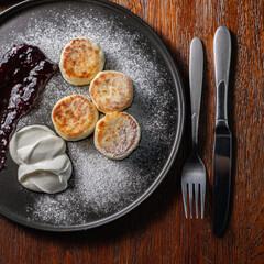 Overhead view of plate with appetizing traditional Russian cheese pancakes with sour cream and berry jam sprinkled with sugar powder on wooden table in cafe 