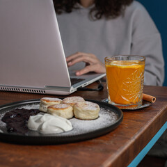 Crop female freelancer working on laptop at table with plate of tasty syrniki dessert and glass of...