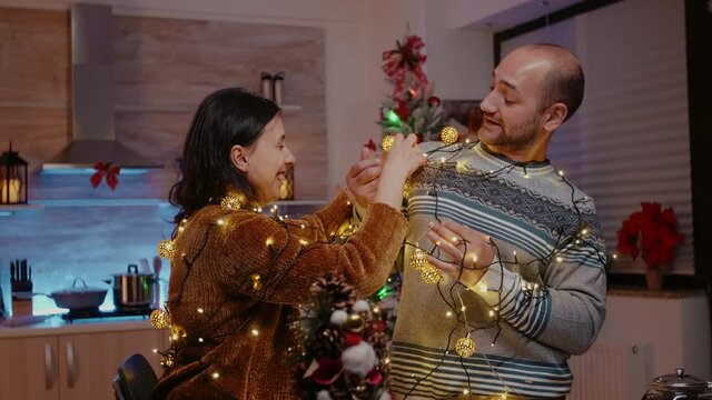 Festive couple trying to untangle garland of twinkle lights while decorating kitchen. Man and woman feeling cheerful and getting tangled in string of christmas lights with illuminated bulbs