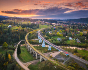 Beautiful old viaduct at sunset in carpathian mountains in autumn in Ukraine. Aerial view of railway bridges, railroad, river, green meadows, trees, houses, hills and colorful sky in fall. Top view