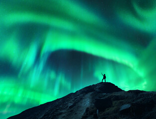 Northern lights and silhouette of standing woman with raised up arms on the mountain. Aurora borealis and happy girl. Sky with stars and green polar lights. Night landscape with aurora. Concept - obrazy, fototapety, plakaty