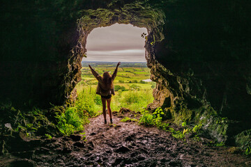Silhouette of a teenager girl standing by a cave entrance her hands up in the air, beautiful nature in the background. Travel, holiday and adventure concept.