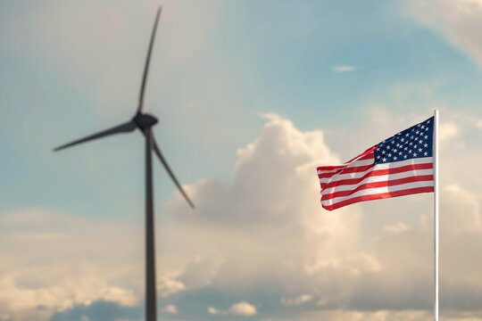 Waving flag of United States of America in focus. Wind power generator turbine and cloudy sky out of focus. Green energy development concept