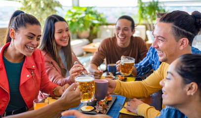 multiethnic group of friends sitting on a table in a bar restaurant making a toast with beers and drinks. diverse people celerating brunch together enjoying happy holidays. lifestyle and joy concept