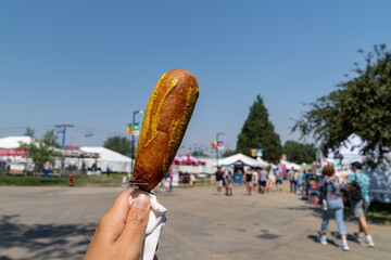 Hand holds up a pronto pup smothered in mustard at the fairgrounds in summer