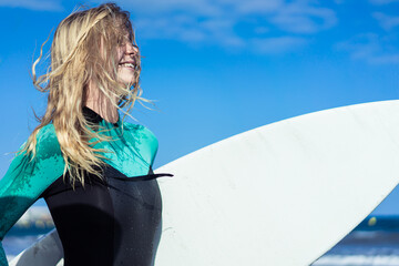 Young sexy girl with long blonde hairs, smiling and jogging with surf board on the beach in a summer day. Background blue sky. Copy space.