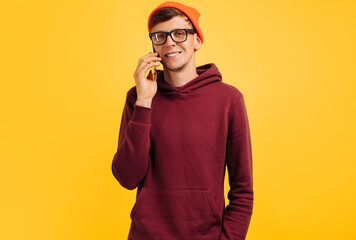 handsome young guy in orange hat glasses and red sweater speaks on phone and laughs while standing on yellow background