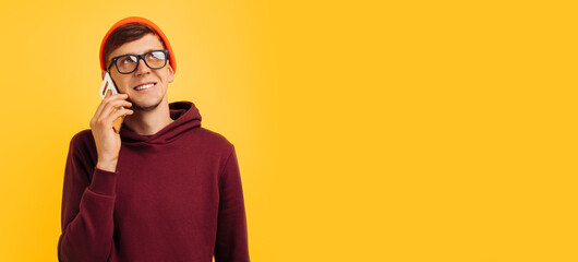 handsome young guy in orange hat glasses and red sweater speaks on phone and laughs while standing on yellow background