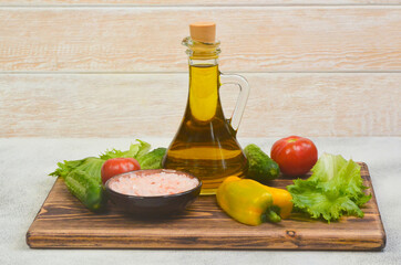 cold-pressed olive oil in a glass bottle with ingredients for cooking summer salad, fresh vegetables tomatoes, cucumbers, bell pepper and green leaf lettuce, pink mineral  himalayan salt seasoning 