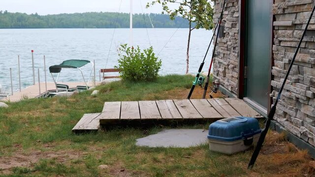 Fishing rods lean against the wall next to the door and wood stoop of a cabin on a tree lined Minnesota lake, with the dock and lake in the distance, in hazy morning light.