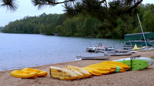 Lake resort beach scene in northern Minnesota with water, sand, trees and various recreational water craft, canoes, kayaks and boats on shore in early morning light.