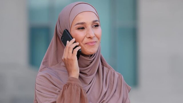 Arabian young happy emotional woman in headscarf standing in city talking cellphone smartphone telephone. Stylish muslim girl wearing hijab speaking on mobile phone answers call chatting conversation