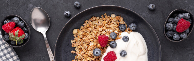 Horizontal banner with homemade granola, yogurt and fresh berries in black ceramic plate on dark stone background. Healthy vegan breakfast