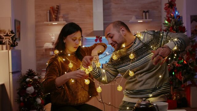 Cheerful Couple Getting Tangled In String Of Twinkle Lights For Christmas Eve Preparations. Man And Woman Laughing While Untangling Knot Of Garland With Lights And Illuminated Bulbs