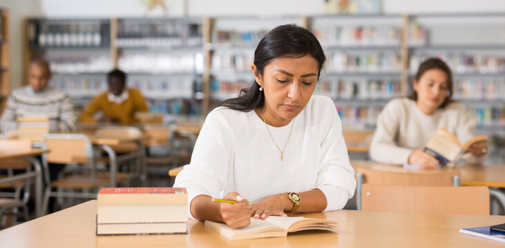 Portrait of focused adult Latina studying in university library, making notes in workbook