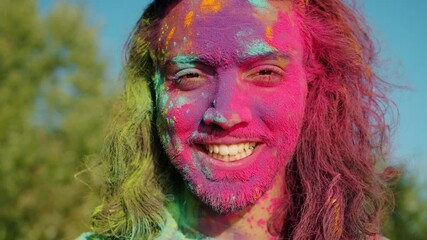 Close-up slow motion portrait of carefree Arab man smiling looking at camera with face covered with gulal paint during Holi celebration outdoors
