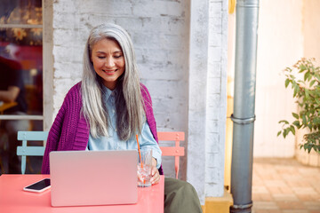 Positive senior Asian woman with glass of water and smartphone looks at laptop screen at coral...