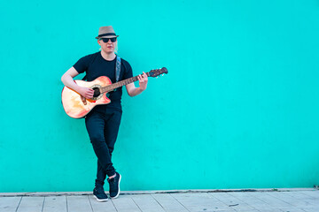 Obraz premium Close-up of albino man with hat playing guitar leaning against a colored wall. Horizontal orientation. Relaxation and inner peace concept.