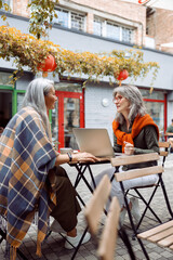 Obraz na płótnie Canvas Mature women business partners with modern laptop discuss project at small table on outdoors cafe terrace on nice autumn day