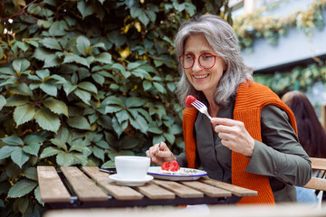 Smiling mature female guest with glasses eats toast with cream and strawberries sitting at table on outdoors cafe terrace on autumn day