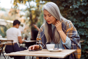 Long haired mature Asian lady reads message on mobile phone sitting at small table on outdoors cafe terrace on autumn day