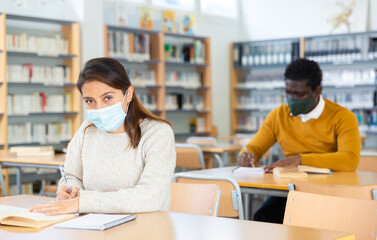 Fototapeta premium Adult female student in face mask working in library, concept of adult education during coronavirus pandemic