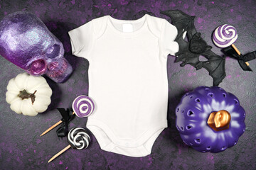 Halloween babywear bay romper onesie bodysuit mockup with purple skull and white pumpkins, lollipops, candy, and bat, on a textured purple background. Product mock up with negative copy space.