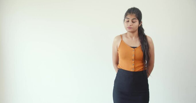 A South Asian female wearing a spaghetti strapped top and black skirt strictly standing against a white wall in 4K