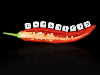 halved red hot chilli pepper with chili seed and the word capsaicin, written with white letter cubes, isolated on black background