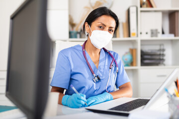 Hispanic woman therapist wearing surgical mask holding consultation in clinic office - obrazy, fototapety, plakaty