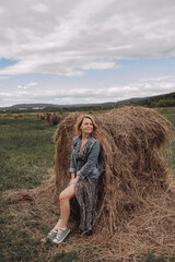 young beautiful woman in long dress, denim jacket, with bare legs in sneakers near large haystack in an empty field. local tourism. enjoying nature. selective focus